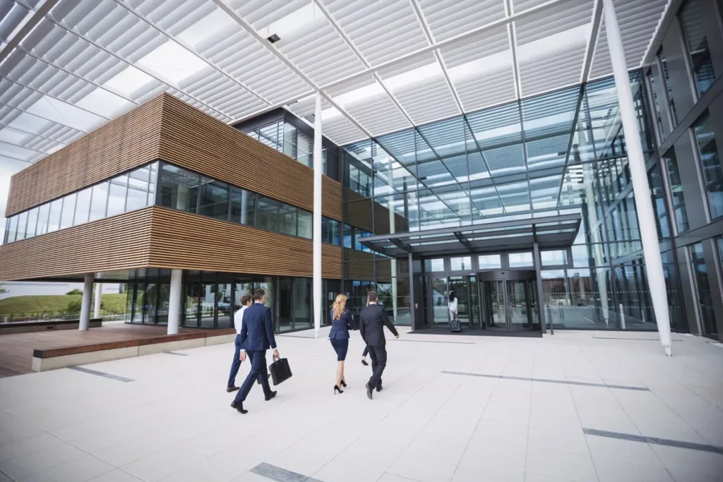 Profissionais em trajes formais caminhando em direção à entrada de um prédio corporativo moderno, com fachada de vidro e elementos de design contemporâneo