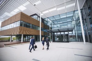 Profissionais em trajes formais caminhando em direção à entrada de um prédio corporativo moderno, com fachada de vidro e elementos de design contemporâneo