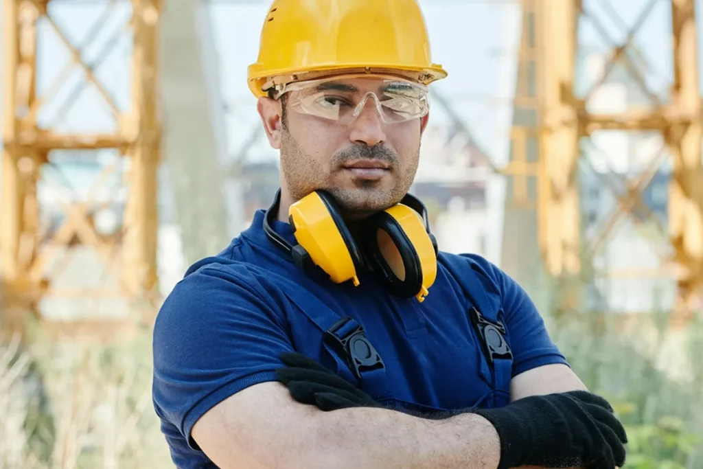 Trabalhador de compliance industrial com capacete amarelo, óculos de proteção e protetor auricular, em um ambiente de construção ao ar livre.