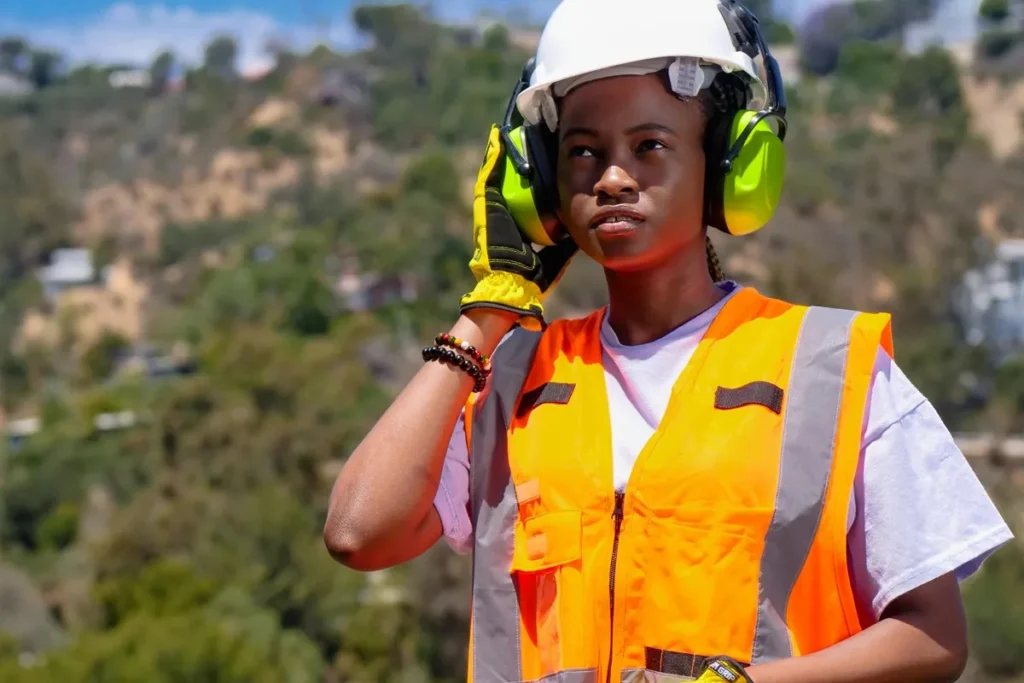 Trabalhadora em ambiente de construção ao ar livre, usando colete laranja refletivo, capacete branco e protetores auriculares verdes, ajustando o equipamento de segurança.