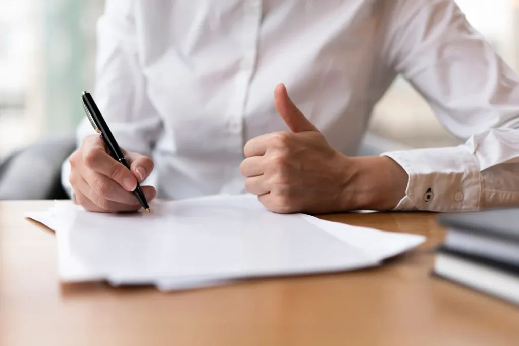 Pessoa assinando um contrato com uma caneta enquanto faz sinal de positivo com a outra mão.