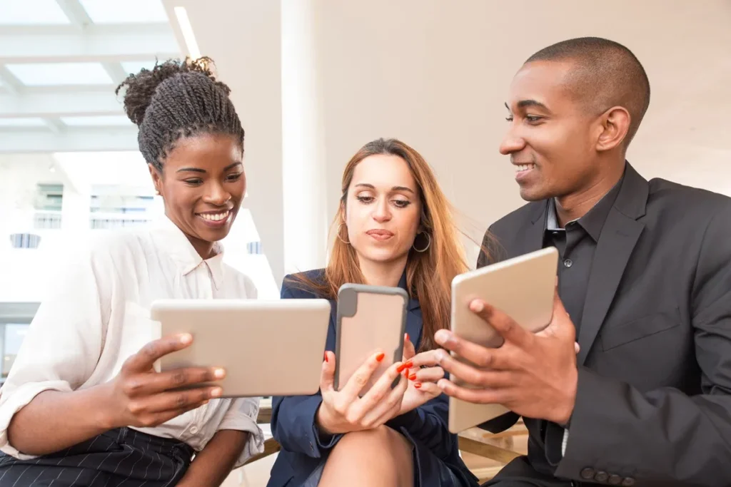 Três profissionais de diferentes origens colaborando e interagindo com dispositivos móveis e tablets em um ambiente corporativo moderno.