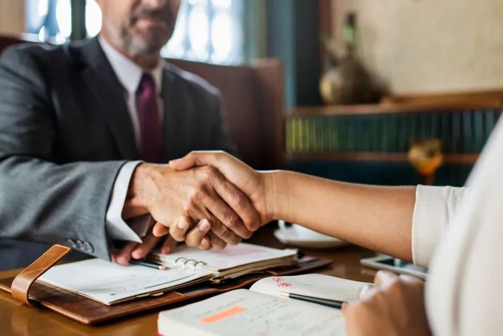 Dois executivos apertando as mãos em um ambiente formal, com cadernos e documentos sobre a mesa, simbolizando o fechamento de um acordo ou parceria.
