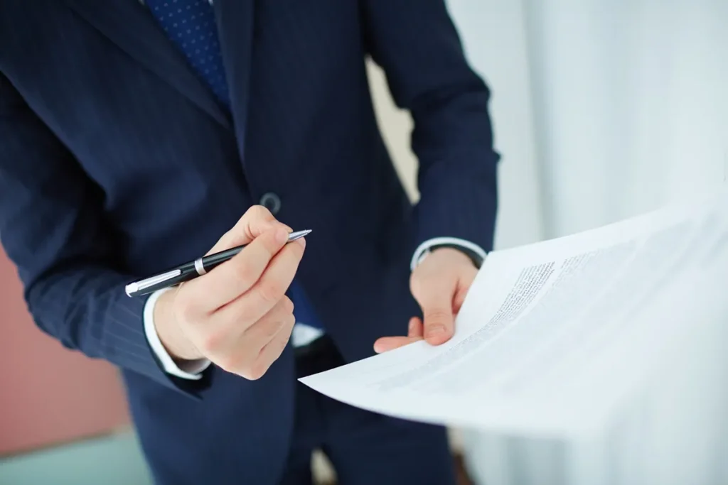 Homem de terno segurando uma caneta e um documento, revisando informações detalhadas em um ambiente corporativo.