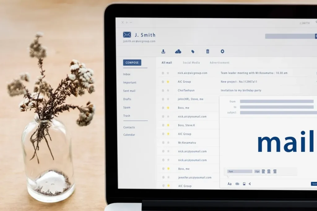 Tela de laptop exibindo uma caixa de entrada de e-mails com organização profissional ao lado de um vaso decorativo com flores secas.