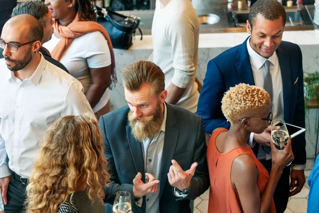 Grupo de profissionais conversando durante um evento corporativo, com destaque para um homem de barba em terno discutindo com colegas ao redor.