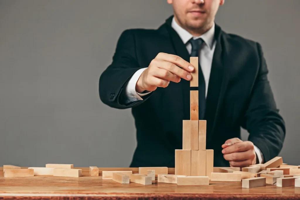 Empresário em traje formal construindo uma torre com blocos de madeira em uma mesa, simbolizando planejamento e estratégia empresarial.