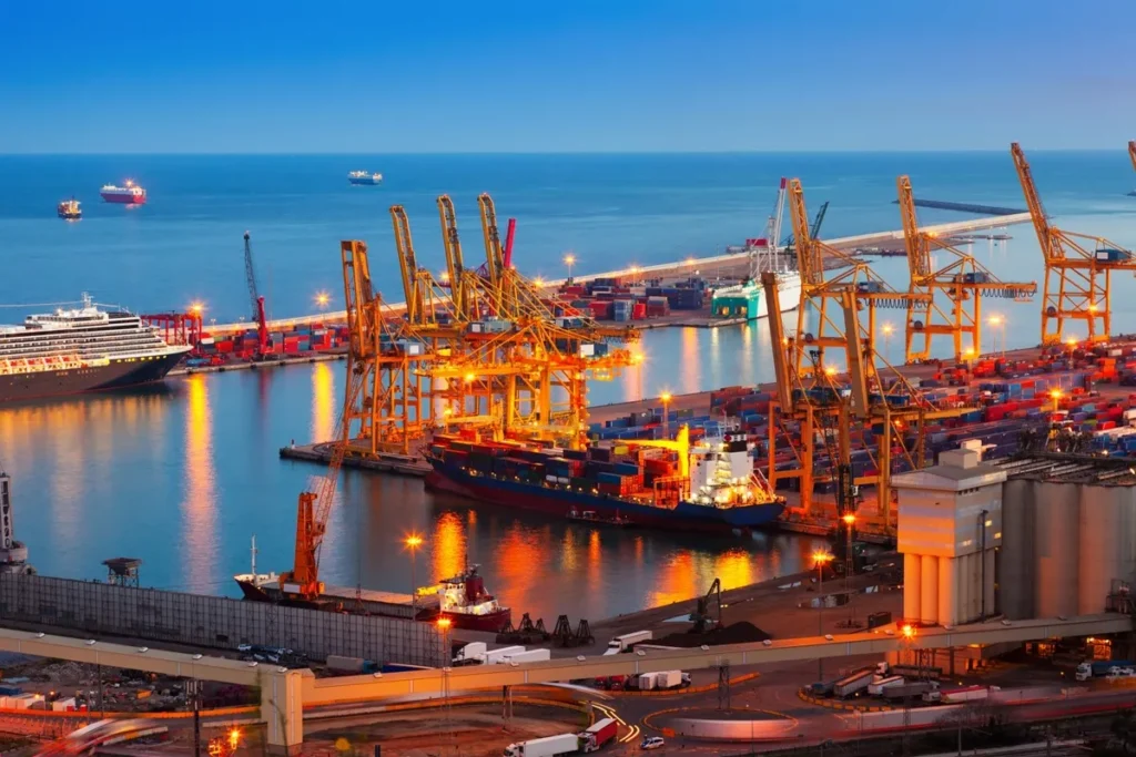 Porto iluminado ao entardecer com guindastes, navios cargueiros e contêineres, mostrando a operação logística de exportação marítima.