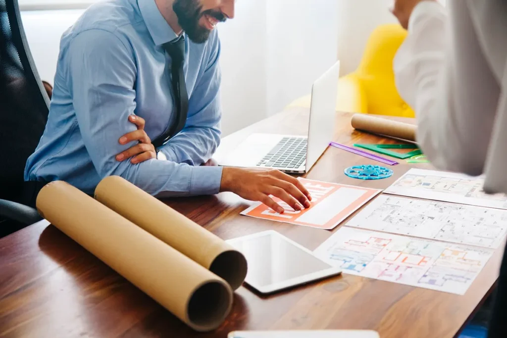 Profissional analisando planos em uma mesa com laptop, desenhos técnicos, rolos de papel e ferramentas de medição.