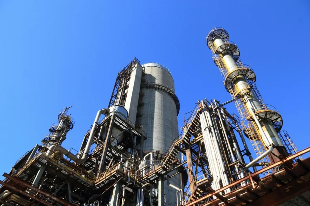 Indústria com estruturas metálicas, tubulações e torres contra um céu azul, representando tecnologia e produção industrial.