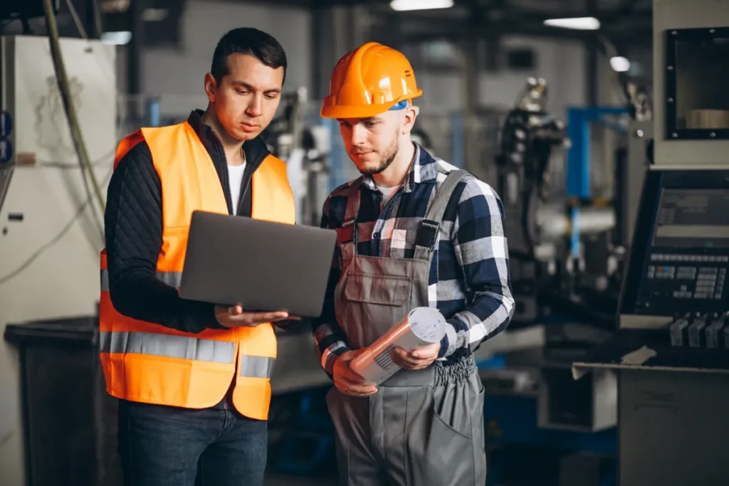 Dois trabalhadores em um ambiente de indústrias, usando um laptop para analisar dados de produção e otimizar processos de fabricação.