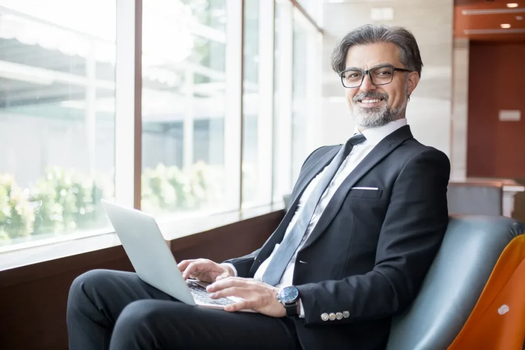 Executivo (CEOs) sorridente com terno e gravata trabalhando em um laptop, sentado próximo a uma grande janela em ambiente corporativo