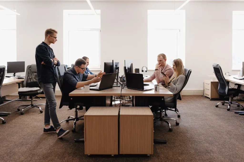 Ambiente corporativo com quatro profissionais trabalhando em um escritório moderno, sentados em estações de trabalho com computadores e um colaborador em pé participando da discussão.