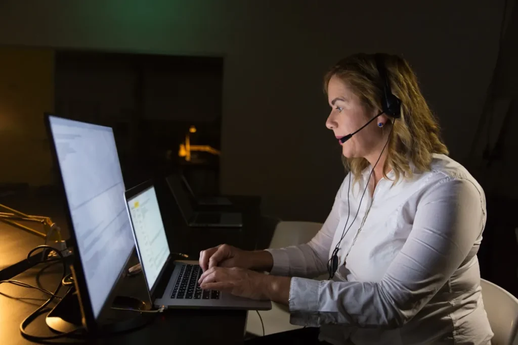 Mulher usando headset trabalhando em um laptop em um ambiente de atendimento ao cliente durante a noite.