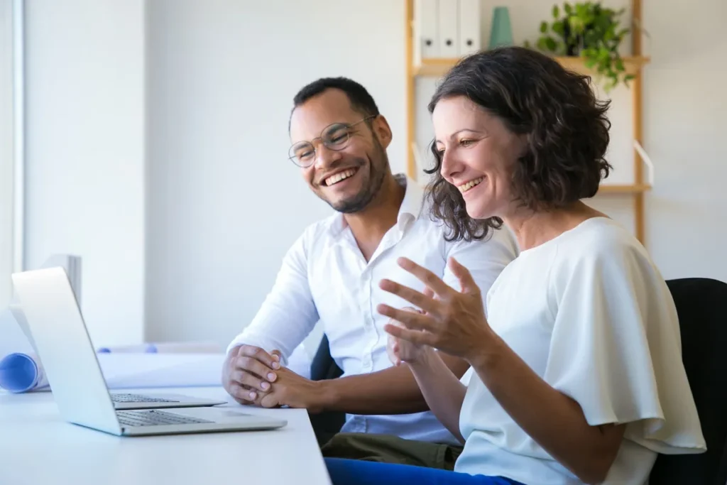 Dois colegas de trabalho sorridentes colaborando em frente a um laptop em um escritório iluminado e moderno.