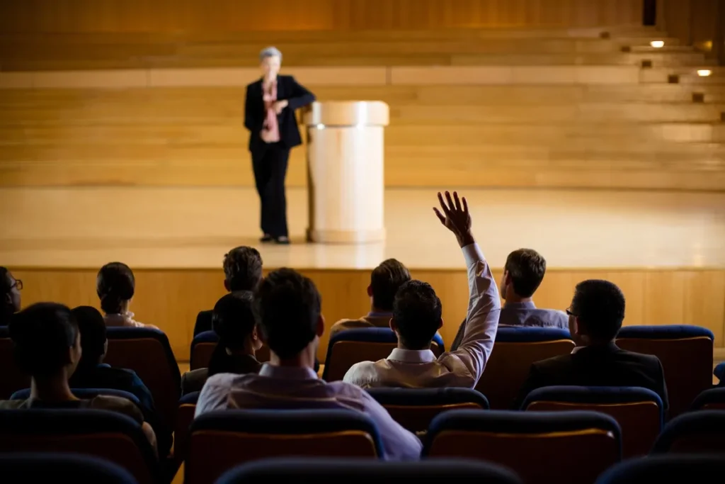 Participante levantando a mão para fazer uma pergunta durante uma conferência, com palestrante em um palco ao fundo.
