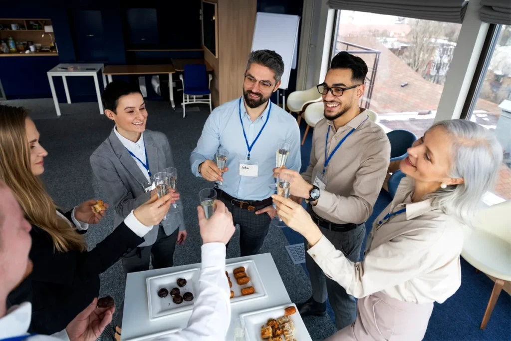 Grupo de profissionais em um evento corporativo, brindando com taças de champanhe e sorrindo, ao redor de uma mesa com aperitivos