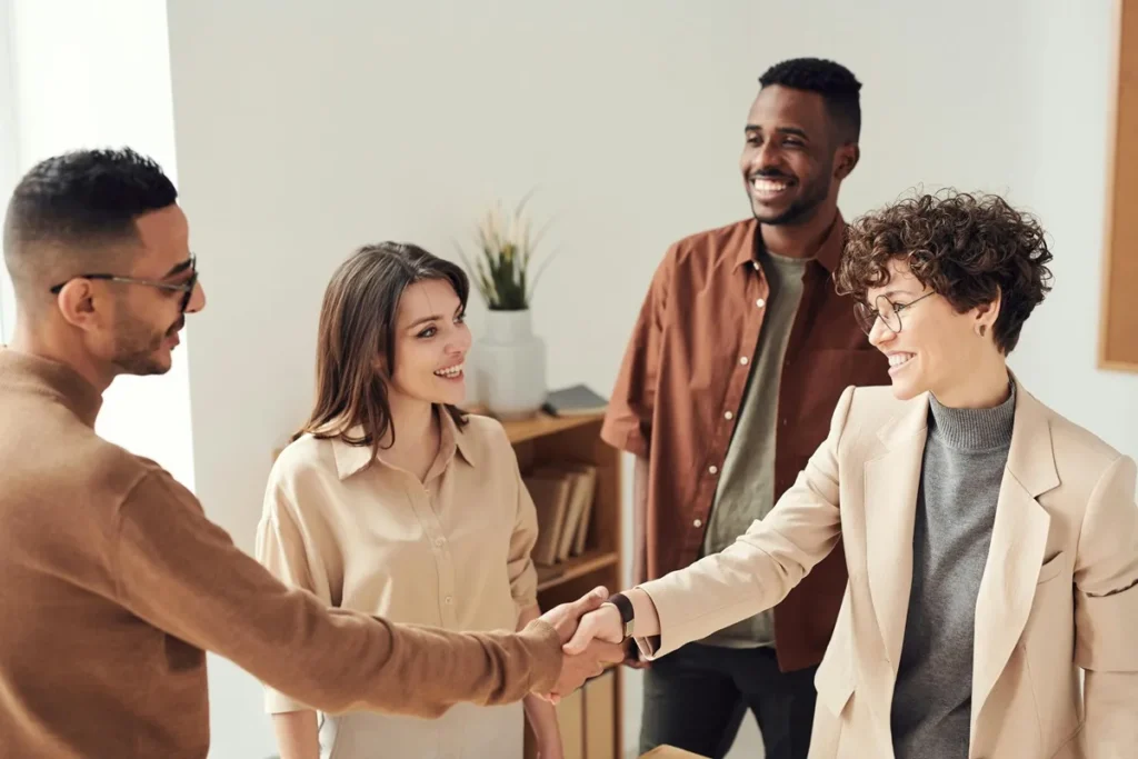Grupo de quatro pessoas sorridentes, duas delas apertando as mãos, simbolizando uma parceria ou negociação bem-sucedida em um ambiente de trabalho.