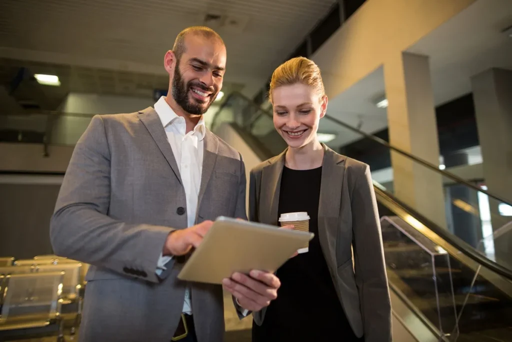 Um homem e uma mulher em trajes formais olhando para um tablet enquanto sorriem, simbolizando trabalho em equipe e colaboração corporativa
