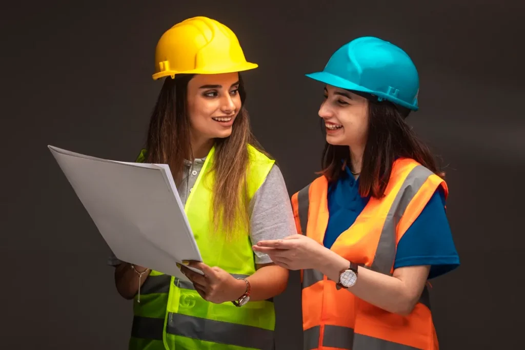 Duas mulheres com capacetes de segurança e coletes refletivos discutindo um projeto em um ambiente interno.
