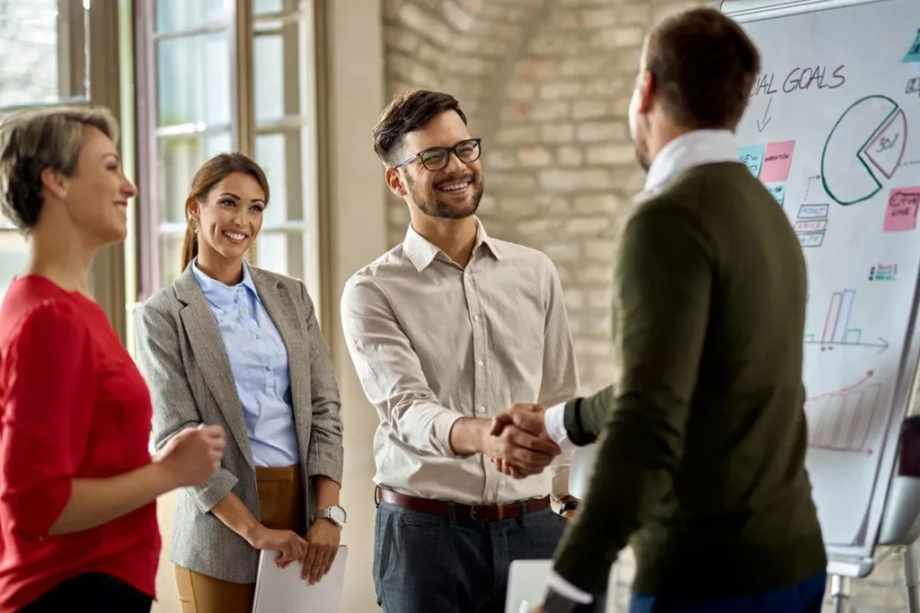 Profissionais em uma reunião corporativa apertando as mãos, simbolizando a formação de uma parceria estratégica, com equipe ao fundo sorrindo.