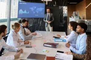 Reunião corporativa com profissional apresentando dados em tela para equipe ao redor de uma mesa