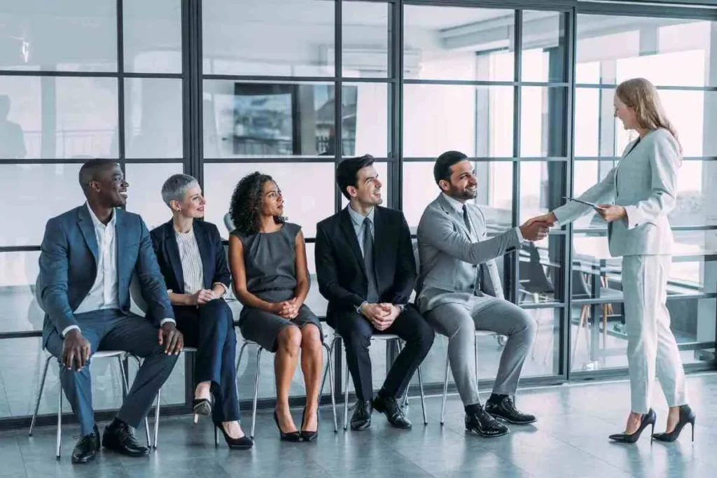 Candidatos sentados em fila para entrevista, enquanto um deles cumprimenta a recrutadora, ilustrando uma etapa do processo seletivo em um escritório.