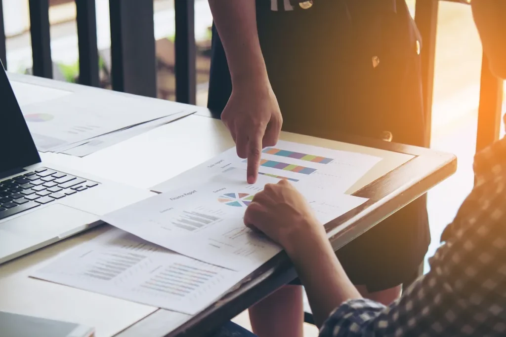 Duas pessoas revisando gráficos e relatórios impressos em uma mesa, com destaque para dados de cores e diagramas.