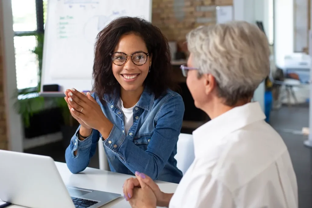Duas mulheres conversando em um ambiente corporativo, com foco na colaboração e troca de ideias em frente a um laptop.