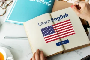 Livro aberto com a frase "Learn English" acompanhada da bandeira dos Estados Unidos, sugerindo um ambiente de estudo de inglês. Uma pessoa segura o livro, e há um caderno azul, uma caneta e uma xícara de chá na mesa.