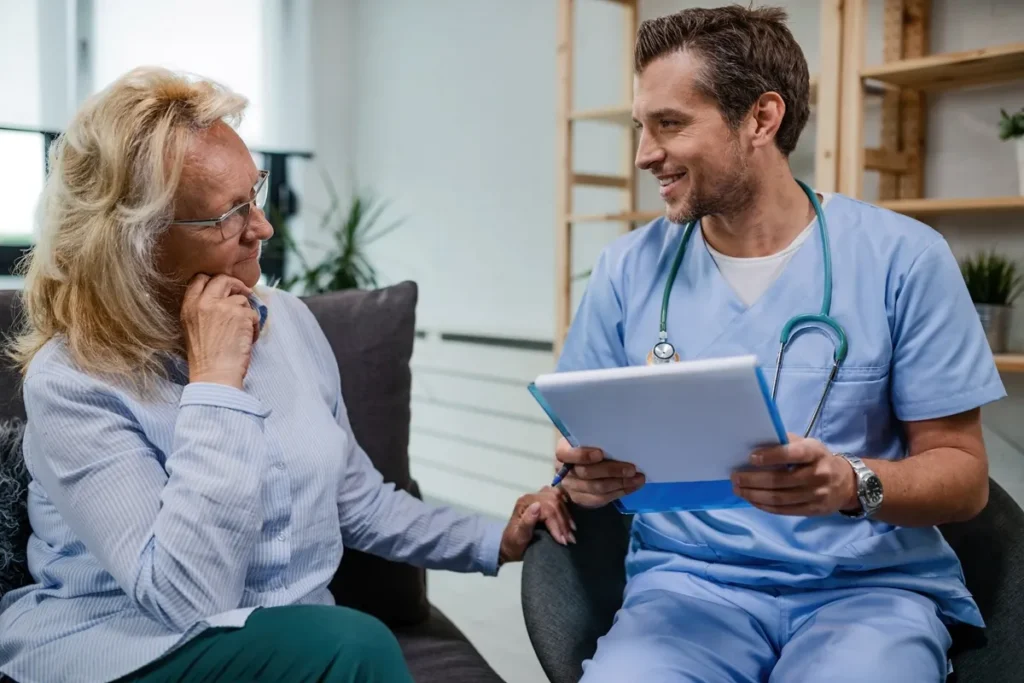 Médico sorridente em uniforme azul e estetoscópio conversando com uma idosa de cabelos loiros e roupa azul-clara em um ambiente aconchegante.