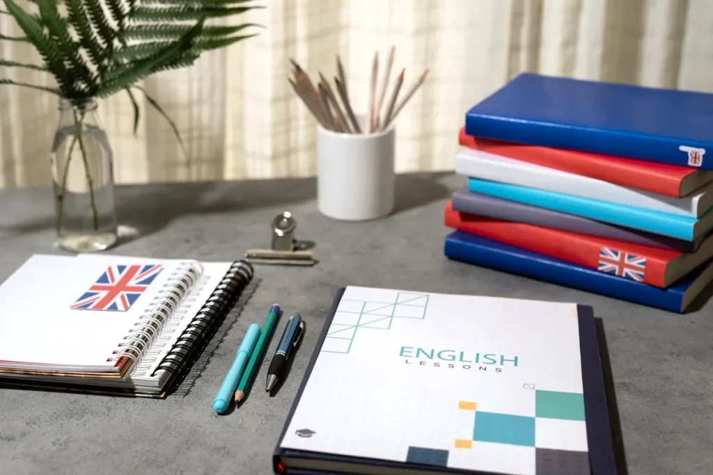 Mesa de estudo com cadernos e livros de inglês, incluindo um com a capa "English Lessons", canetas e um caderno espiral com a bandeira do Reino Unido, sugerindo um ambiente de aprendizado do idioma.