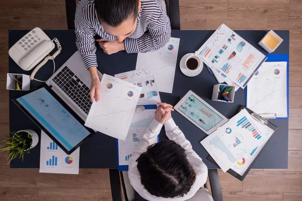 Vista aérea de uma mesa de escritório com relatórios de dados, gráficos e dispositivos digitais, onde duas pessoas discutem estratégias para otimizar a conversão em negócios.