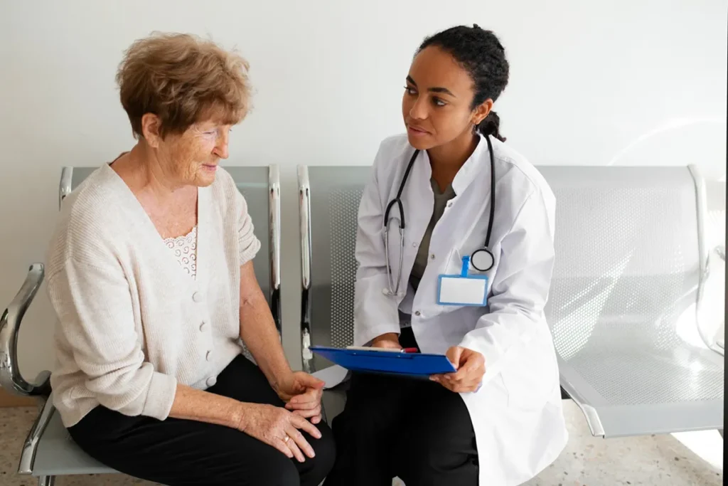 Médica jovem com jaleco branco e estetoscópio conversando de forma atenciosa com uma idosa sentada em um banco de espera.