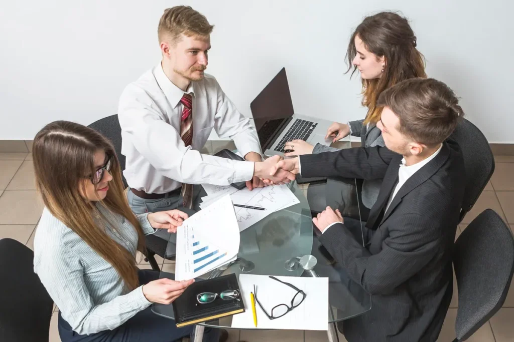 Profissionais em uma reunião de negócios formal negociando em inglês, analisando gráficos financeiros e fechando um acordo com um aperto de mãos.