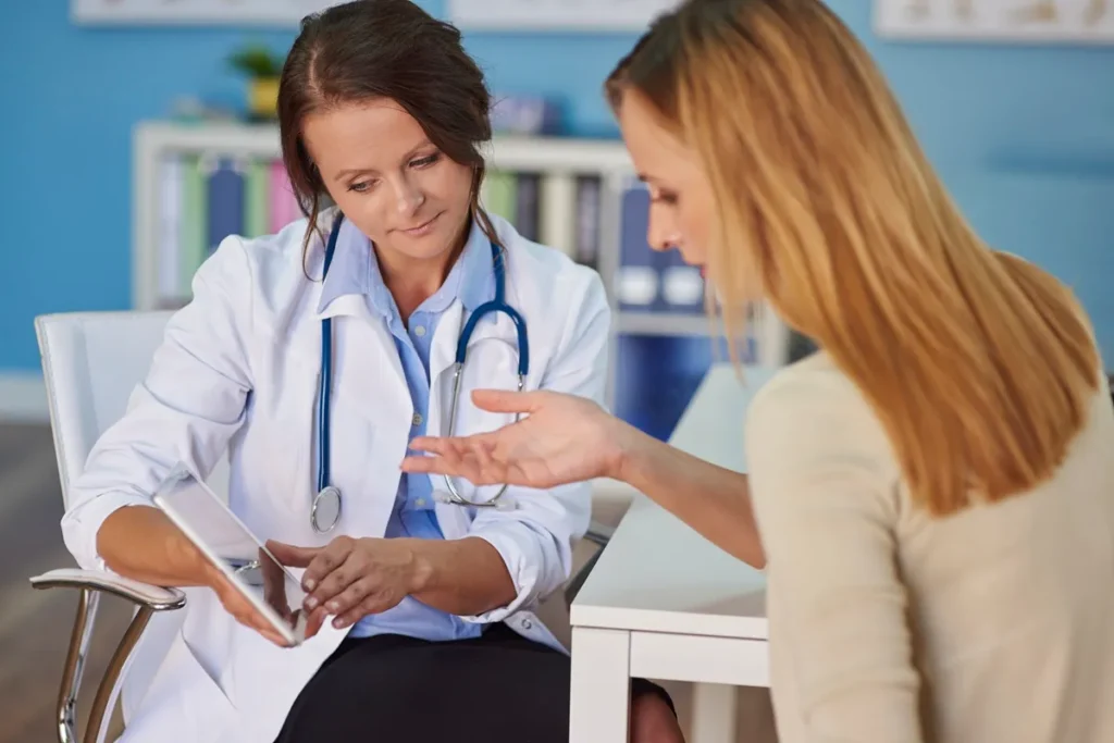 Médica atenciosa com jaleco branco e estetoscópio utilizando um tablet para mostrar informações a uma paciente loira em um consultório moderno.