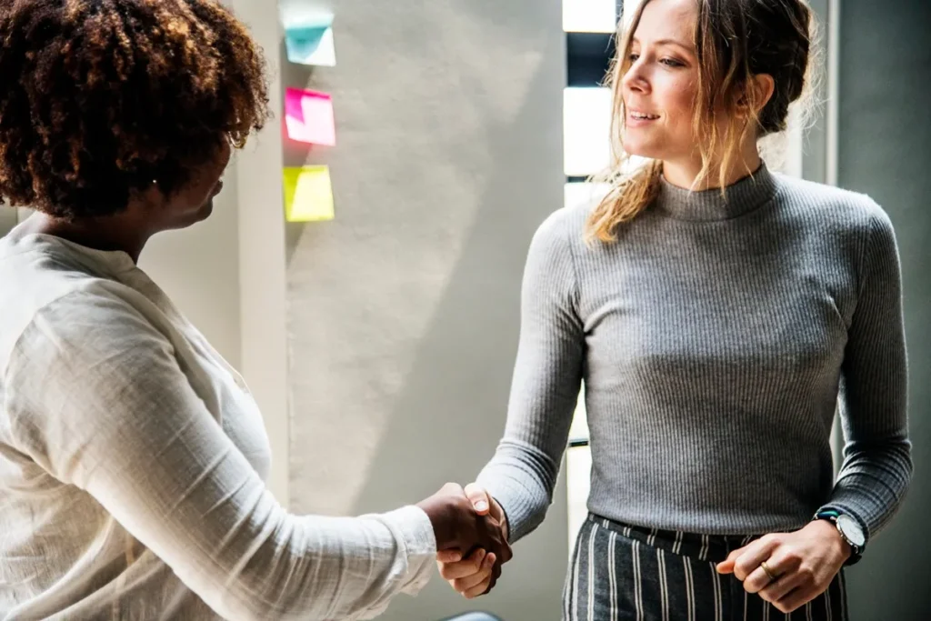 Duas mulheres apertando as mãos em um ambiente profissional, simbolizando um acordo ou networking, com post-its coloridos ao fundo.