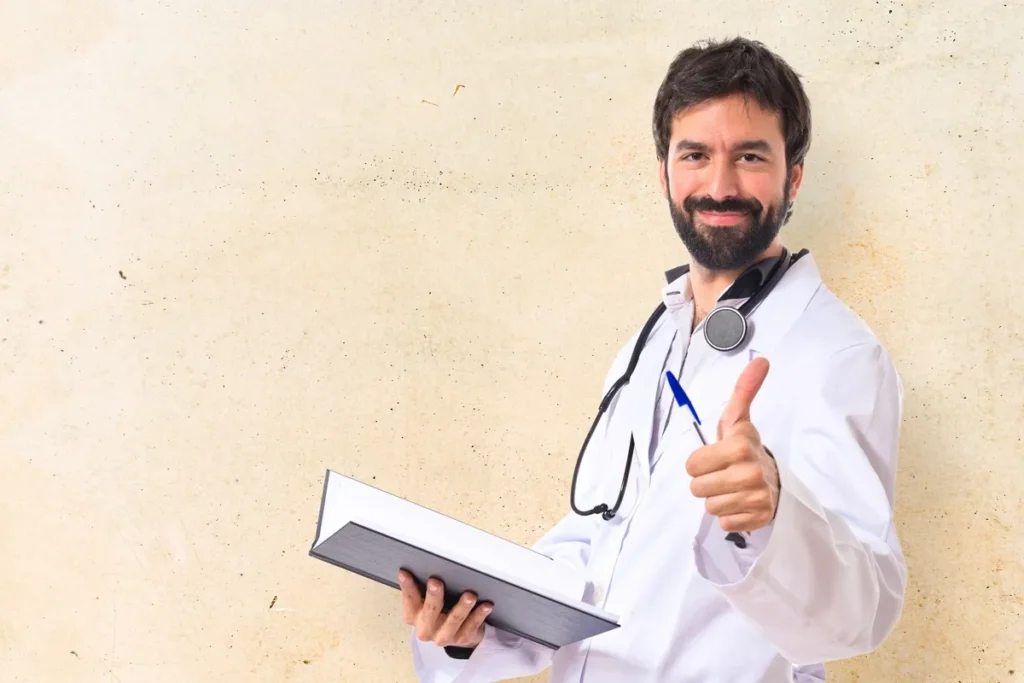 Médico sorridente com barba, vestindo jaleco branco e estetoscópio, segurando uma prancheta e fazendo um sinal de positivo.