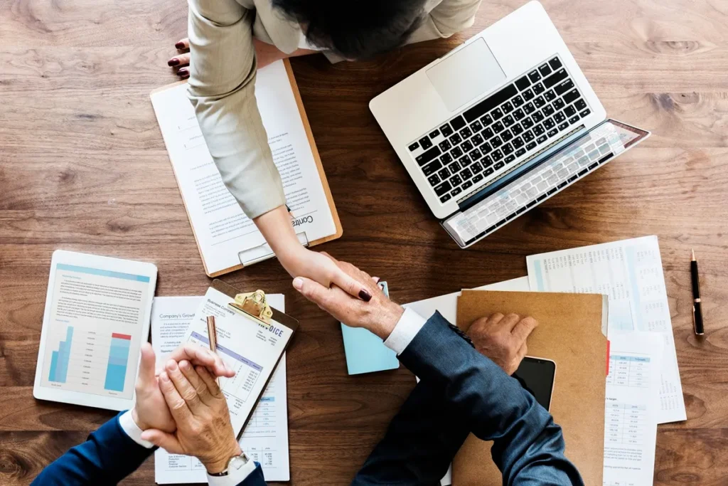 Vista superior de uma negociação de negócios, onde duas pessoas apertam as mãos sobre uma mesa repleta de documentos financeiros, um laptop e relatórios.