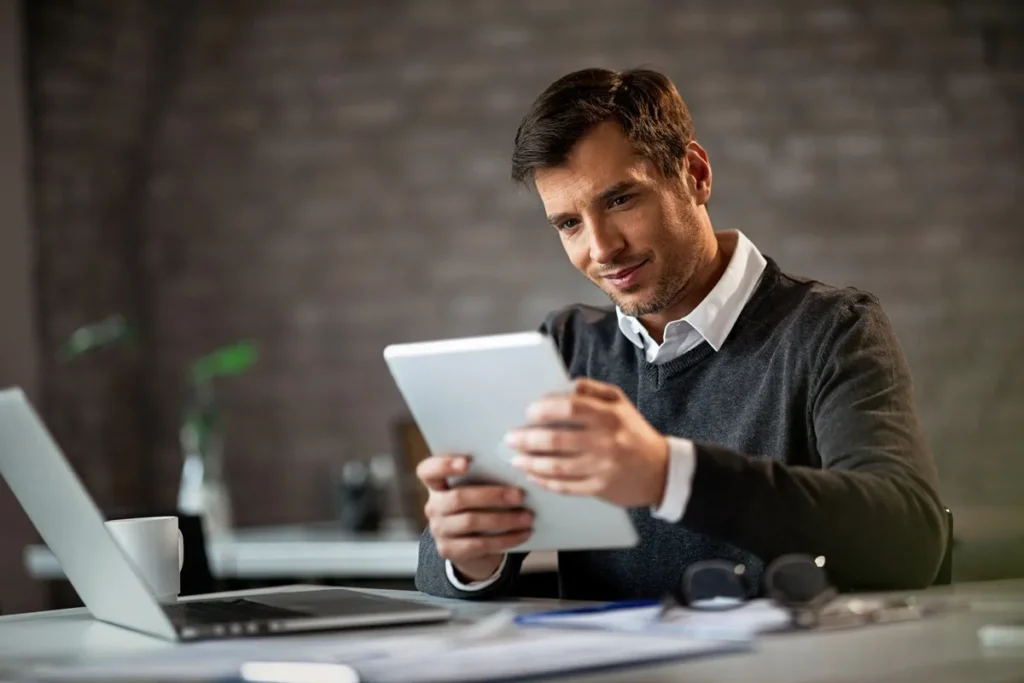 Homem sorridente segurando um tablet enquanto trabalha em um ambiente corporativo, com um laptop e documentos sobre a mesa.