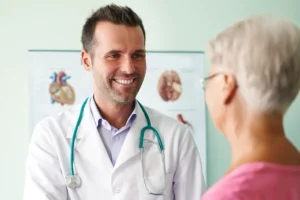 Médico sorridente com jaleco branco e estetoscópio conversando com uma paciente idosa de cabelos grisalhos em um consultório médico.