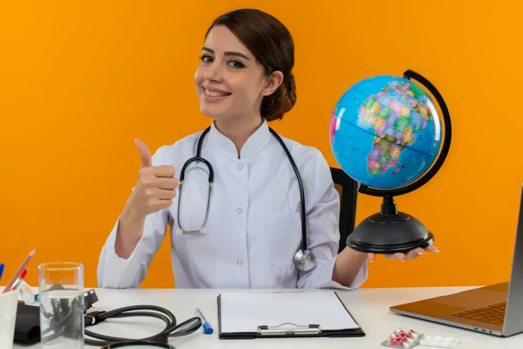 Médica sorridente com jaleco branco e estetoscópio, segurando um globo terrestre e fazendo um sinal de positivo, em fundo laranja.