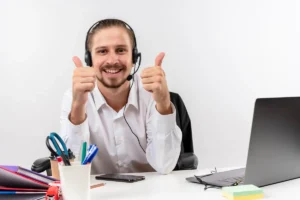 Homem sorridente com headset e camisa social branca, mostrando sinal positivo com as mãos em um ambiente de escritório, representando suporte ao cliente.
