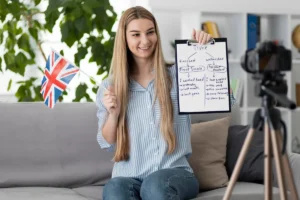 Mulher jovem com cabelos longos e loiros, vestindo uma camisa listrada azul e branca, segurando uma bandeira do Reino Unido em uma mão e um quadro branco na outra. O quadro apresenta uma explicação gramatical sobre a diferença entre "Past Simple" e "Present Perfect" em inglês. Ela está sentada em um sofá e sorri para a câmera, que está posicionada em um tripé à sua frente, gravando a aula. Ao fundo, há uma estante com livros e objetos decorativos, além de plantas verdes que tornam o ambiente aconchegante.