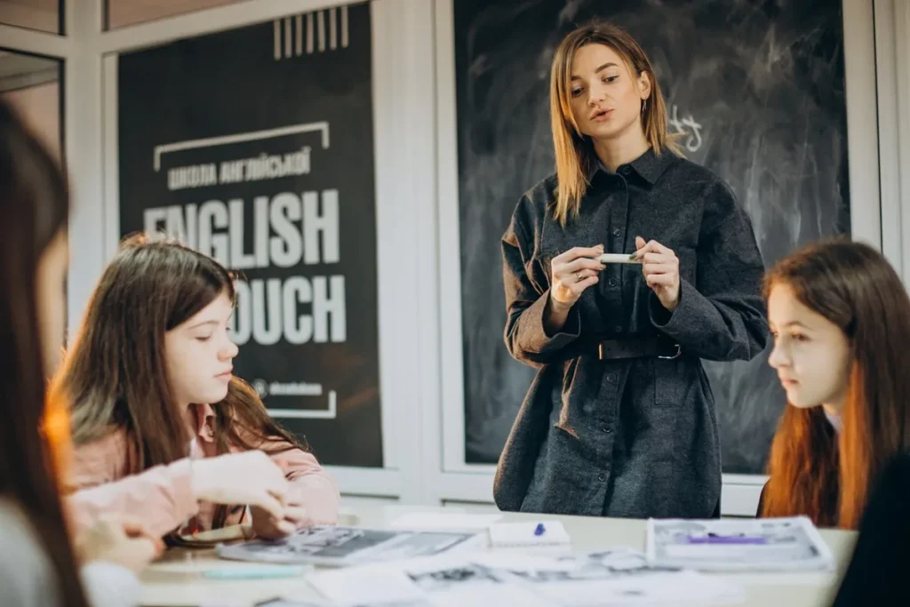 Professora ensinando inglês para crianças em uma sala de aula com quadro negro ao fundo e materiais de estudo sobre a mesa.
