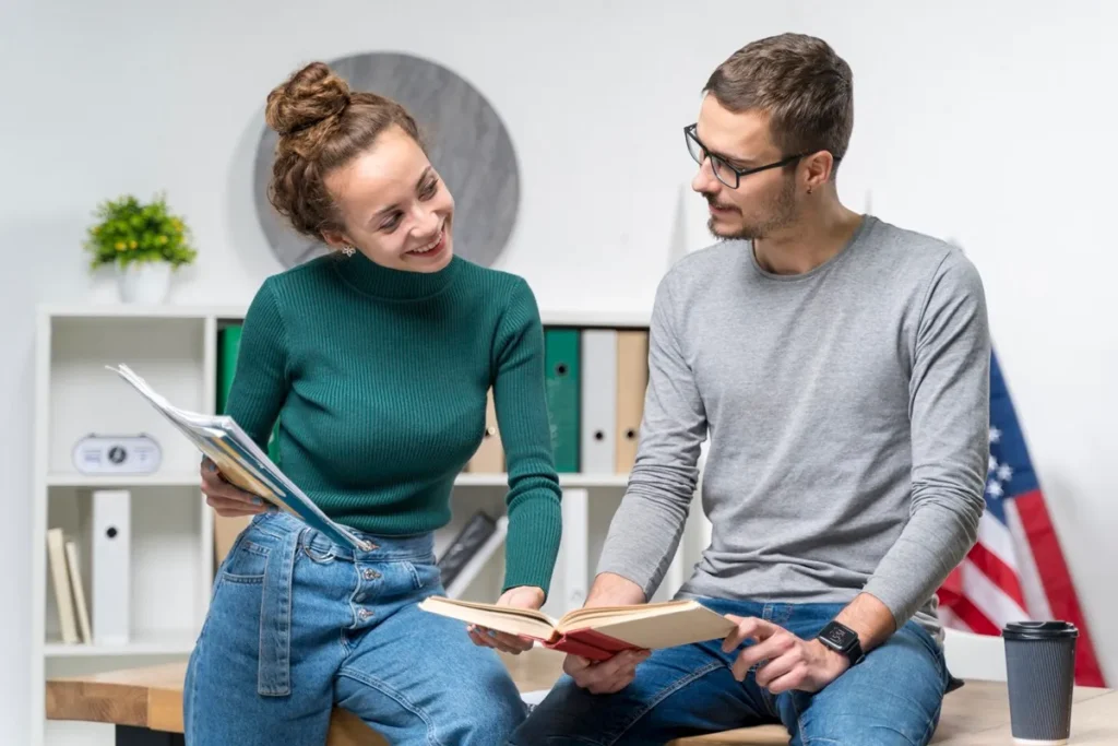 Jovem casal estudando juntos em um ambiente moderno, segurando livros e anotações, com uma bandeira dos EUA ao fundo, sugerindo aprendizado da língua inglesa.