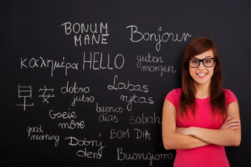 Mulher sorridente de óculos e camiseta rosa em frente a um quadro negro com diversas saudações escritas em diferentes idiomas, incluindo inglês, francês, alemão e chinês.