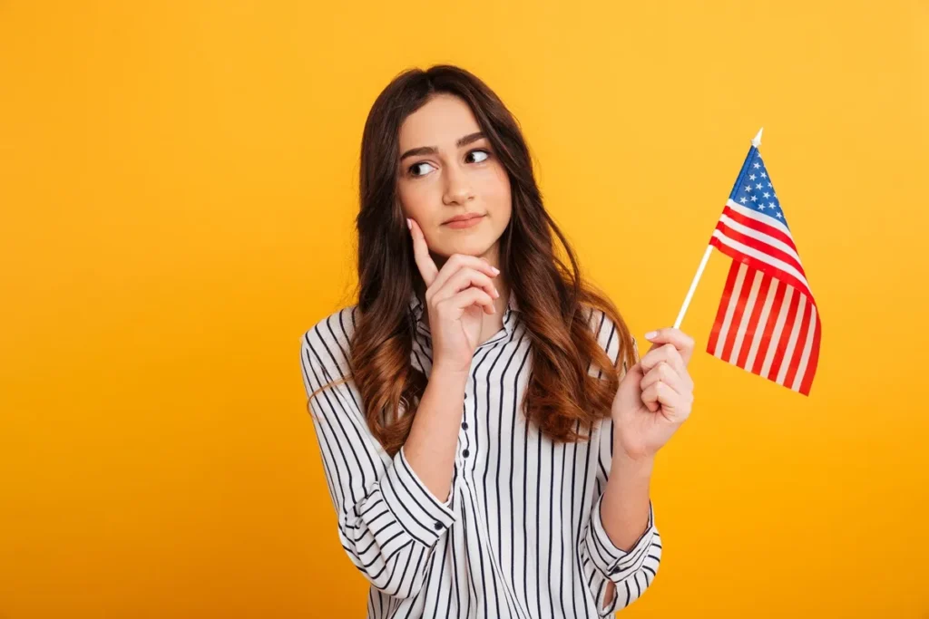Jovem mulher com expressão pensativa segurando a bandeira dos Estados Unidos diante de um fundo amarelo vibrante.