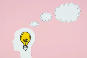 Ilustração de um perfil humano com uma lâmpada amarela dentro da cabeça, representando criatividade e ideias, com balões de pensamento saindo para um fundo rosa.
