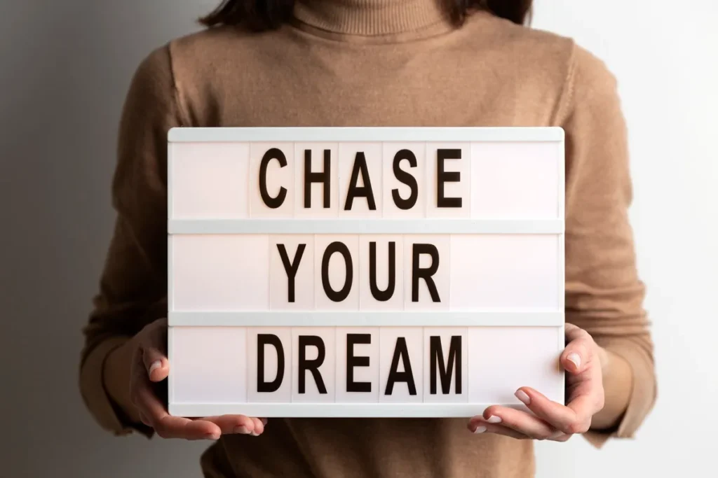 Mulher segurando um quadro iluminado com a frase "Chase Your Dream" escrita em letras pretas, incentivando a busca pelos sonhos.